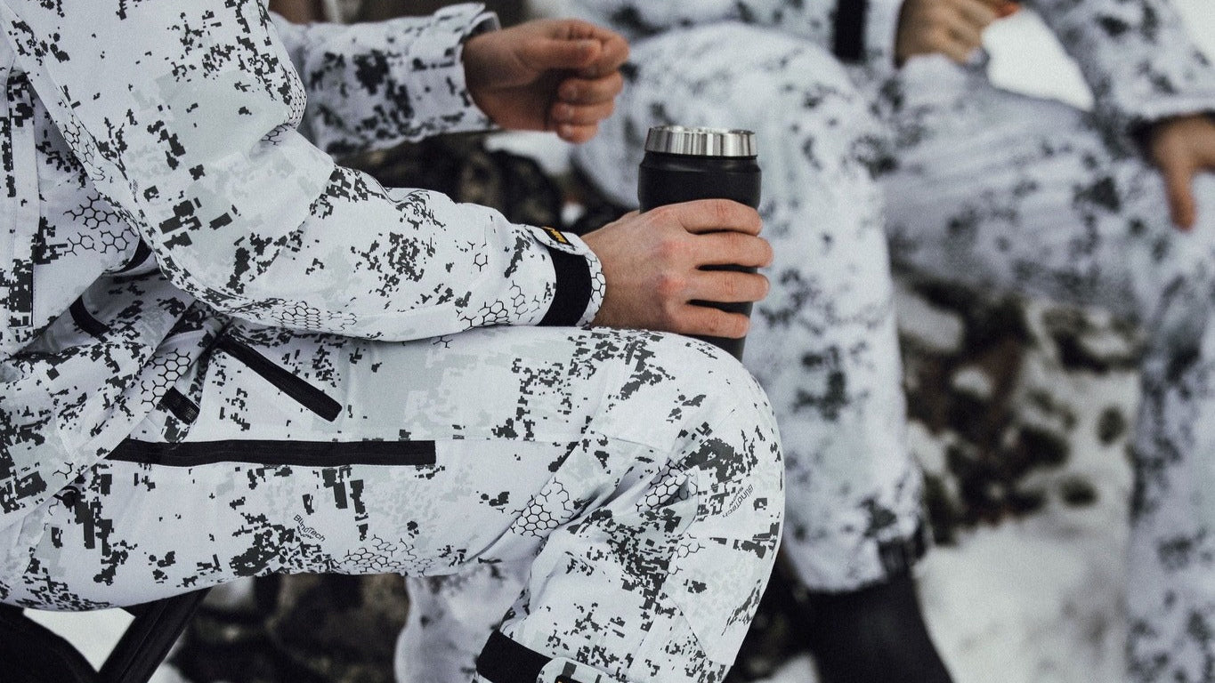 Person in BlindTech snow camouflage gear sitting in the snow holding a thermos.